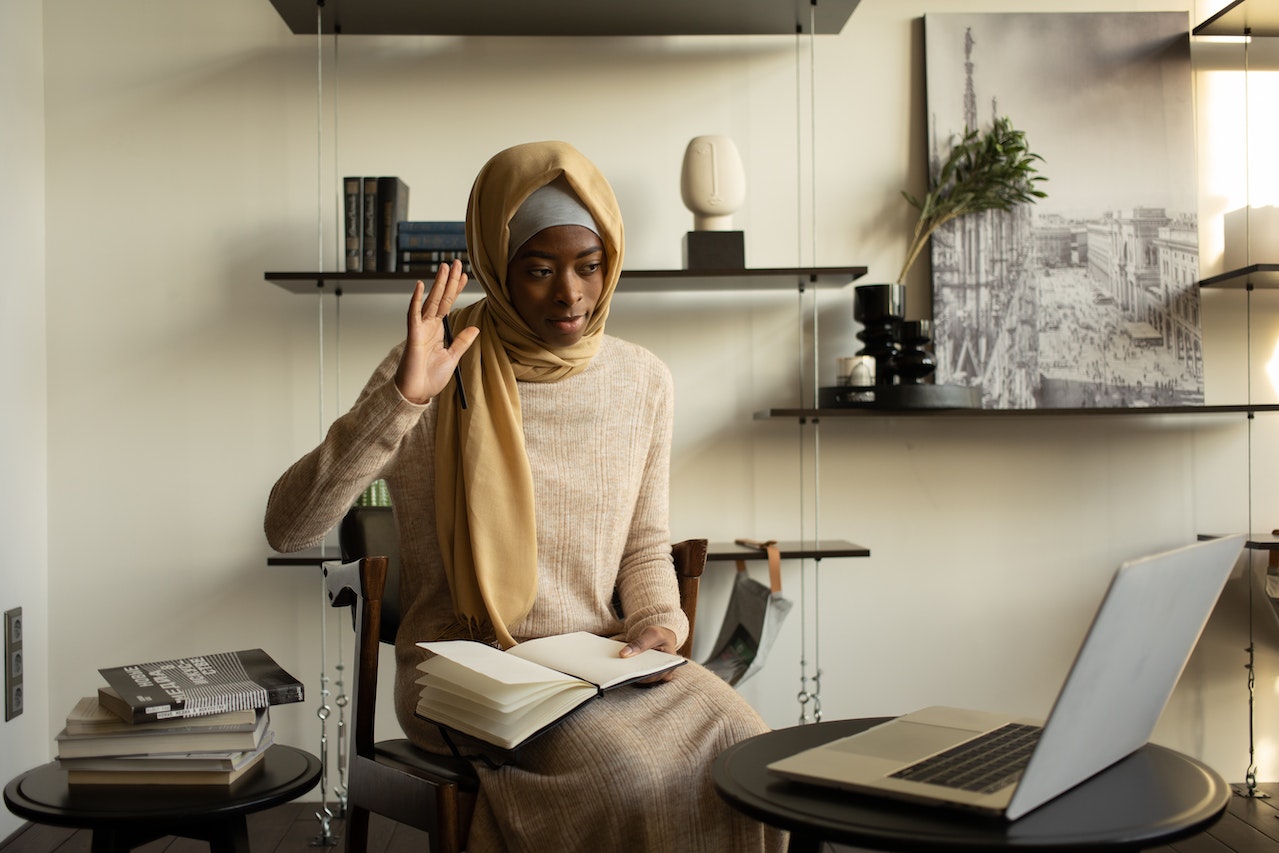 Black-muslim-woman-with-notebook-raising-hand-to-speak-at-videocall-via-laptop-participative-leadership