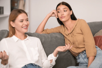 Jealous Lady Listening To Her Cheerful Friend Talking about her Life Sitting On Couch
