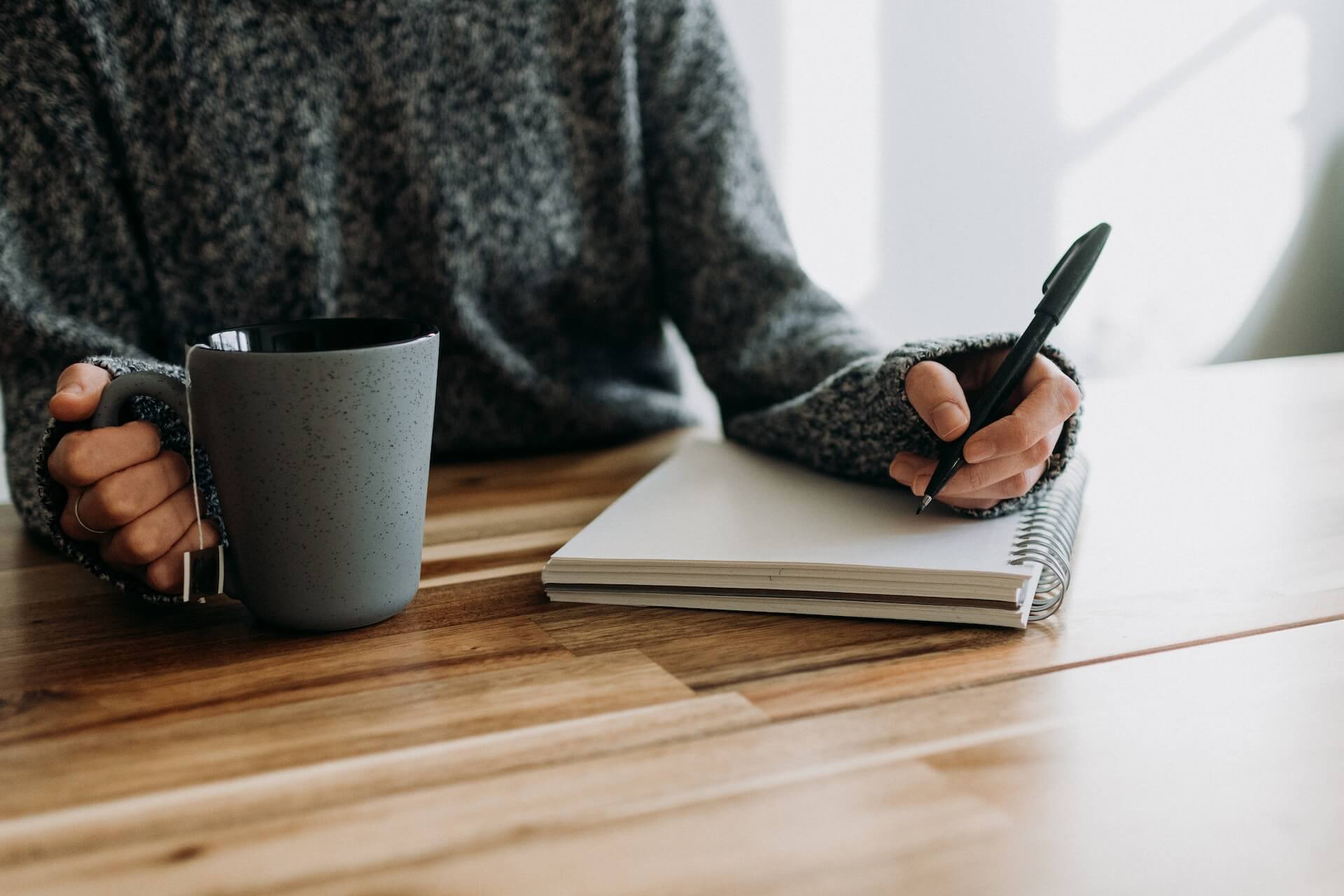 hands of person writing in spiral notebook and hold a mug of tea