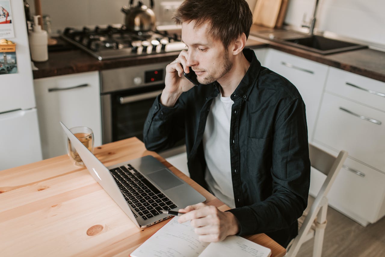 male-freelancer-making-phone-call-and-watching-laptop-in-kitchen