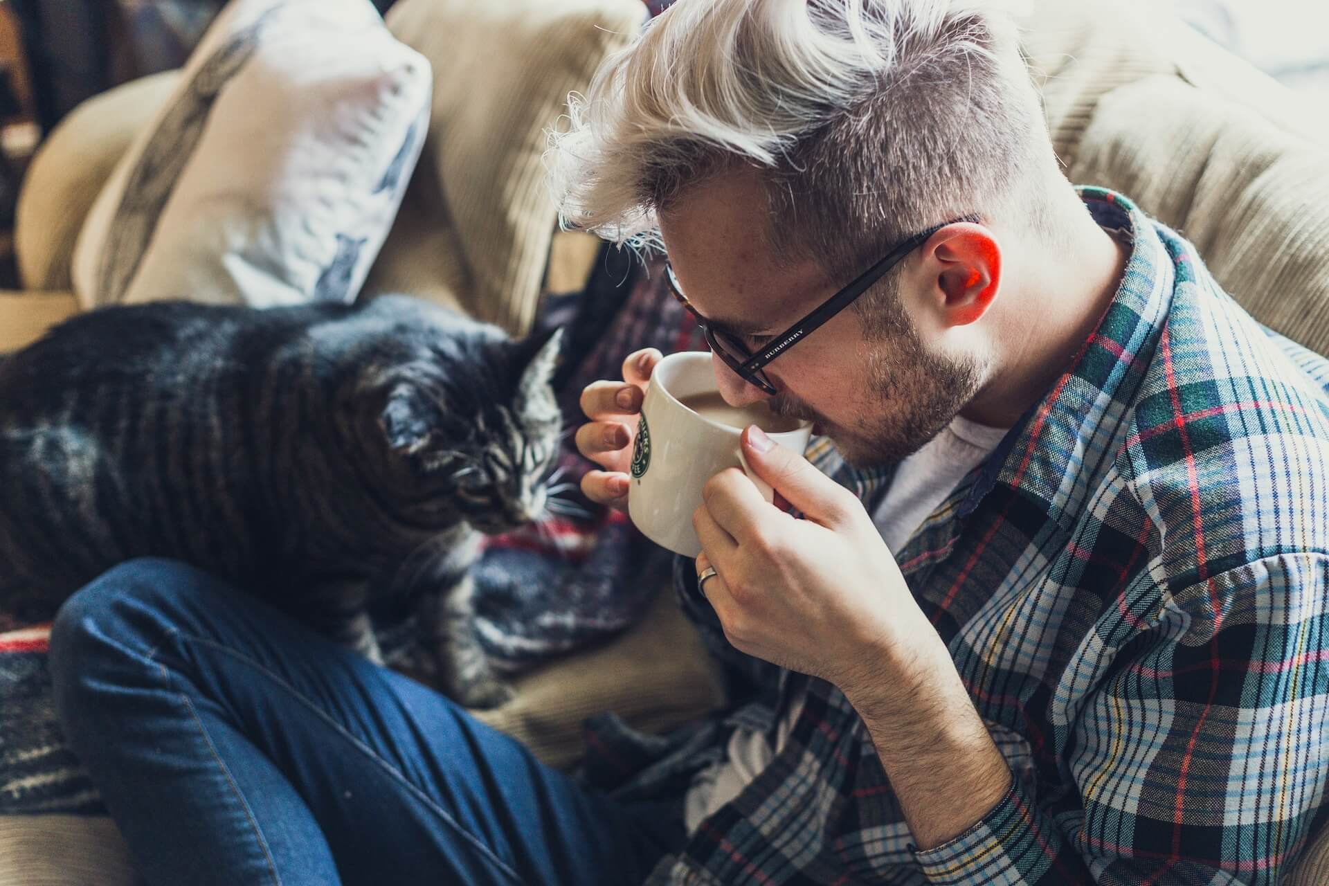 man with glasses drinking from mug with grey cat on sofa