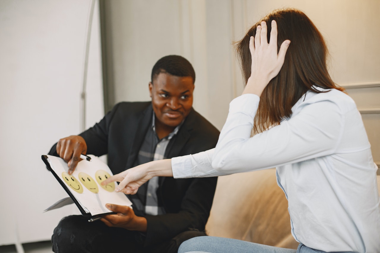 woman-talking-to-man-pointing-to-smiley-face-on-printed-paper-assertive-vs-agressive